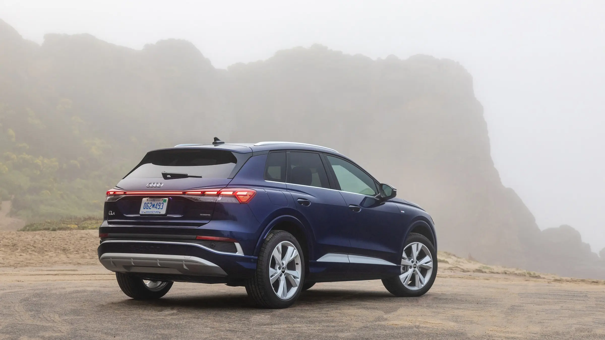 A dark blue Audi Q4 e-tron parked on sand below a fog-covered coastal cliff, seen from the rear three-quarter