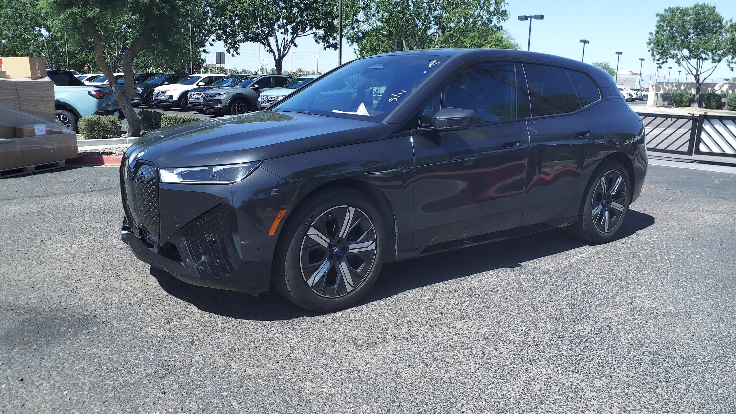 A dark grey BMW iX parked on a sunny dealership lot with a row of pickups and palm-lined streets behind it
