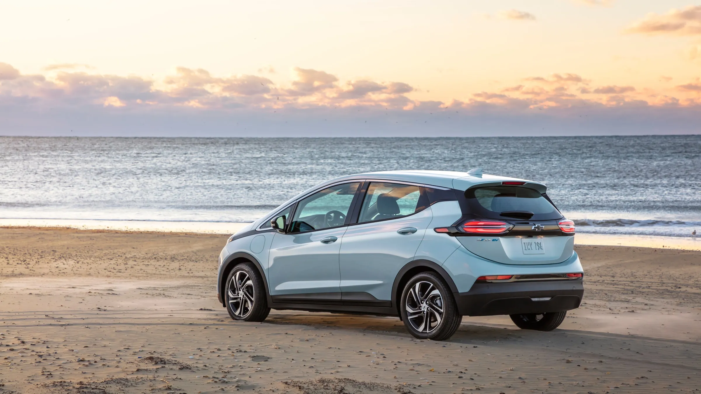 A pale blue Chevrolet Bolt EV parked on a beach at sunset, seen from the rear with its Bolt EV badge and plate showing