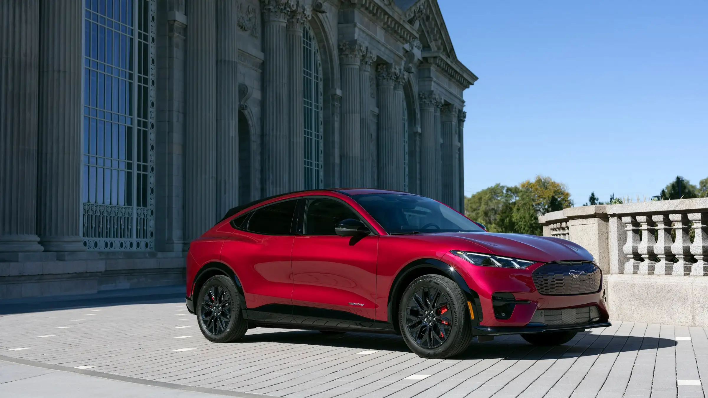 A red Ford Mustang Mach-E parked on a paved plaza beside a tall columned stone building