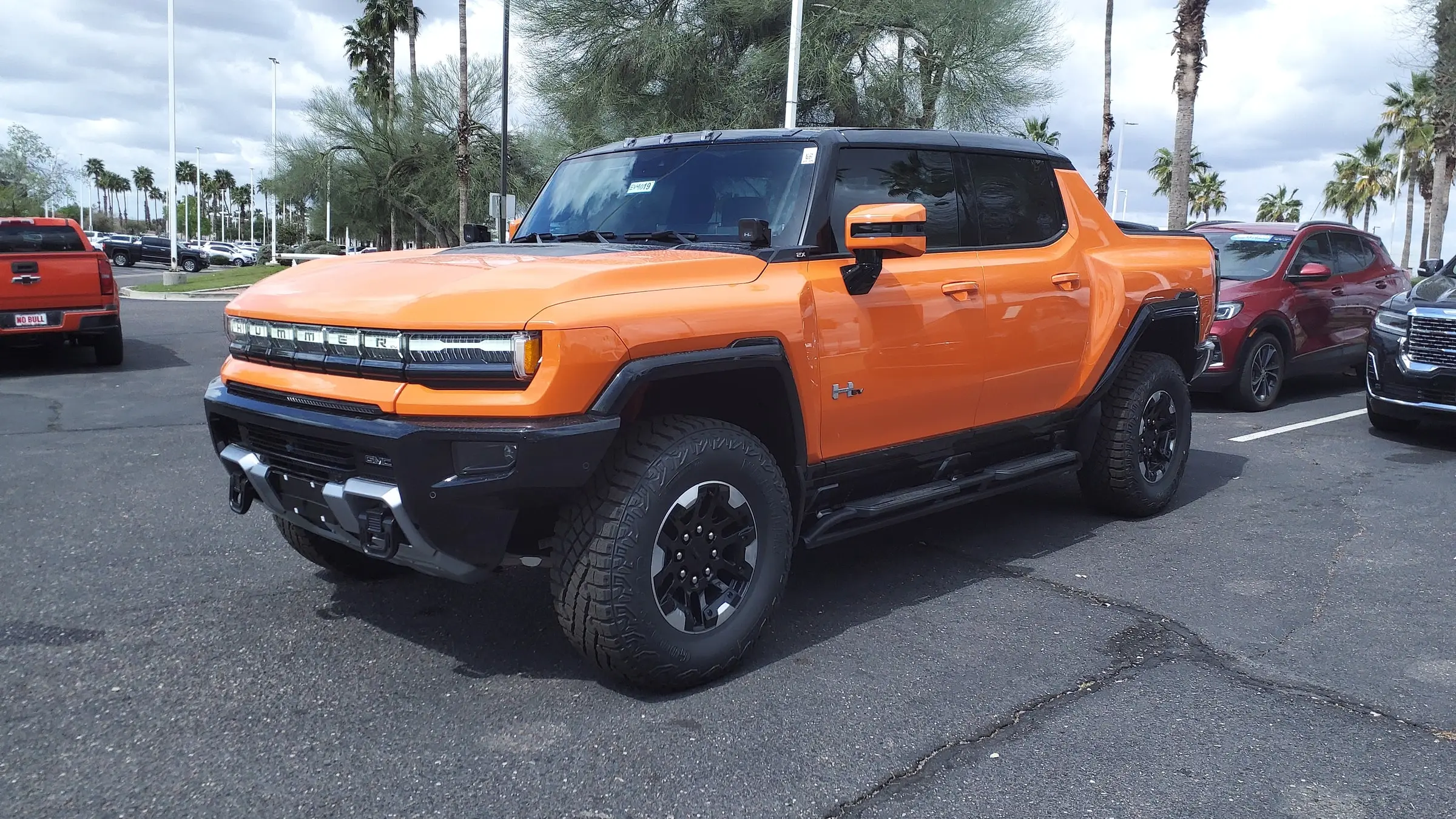 An orange GMC Hummer EV Pickup parked on a dealership lot under palm trees and a cloudy sky