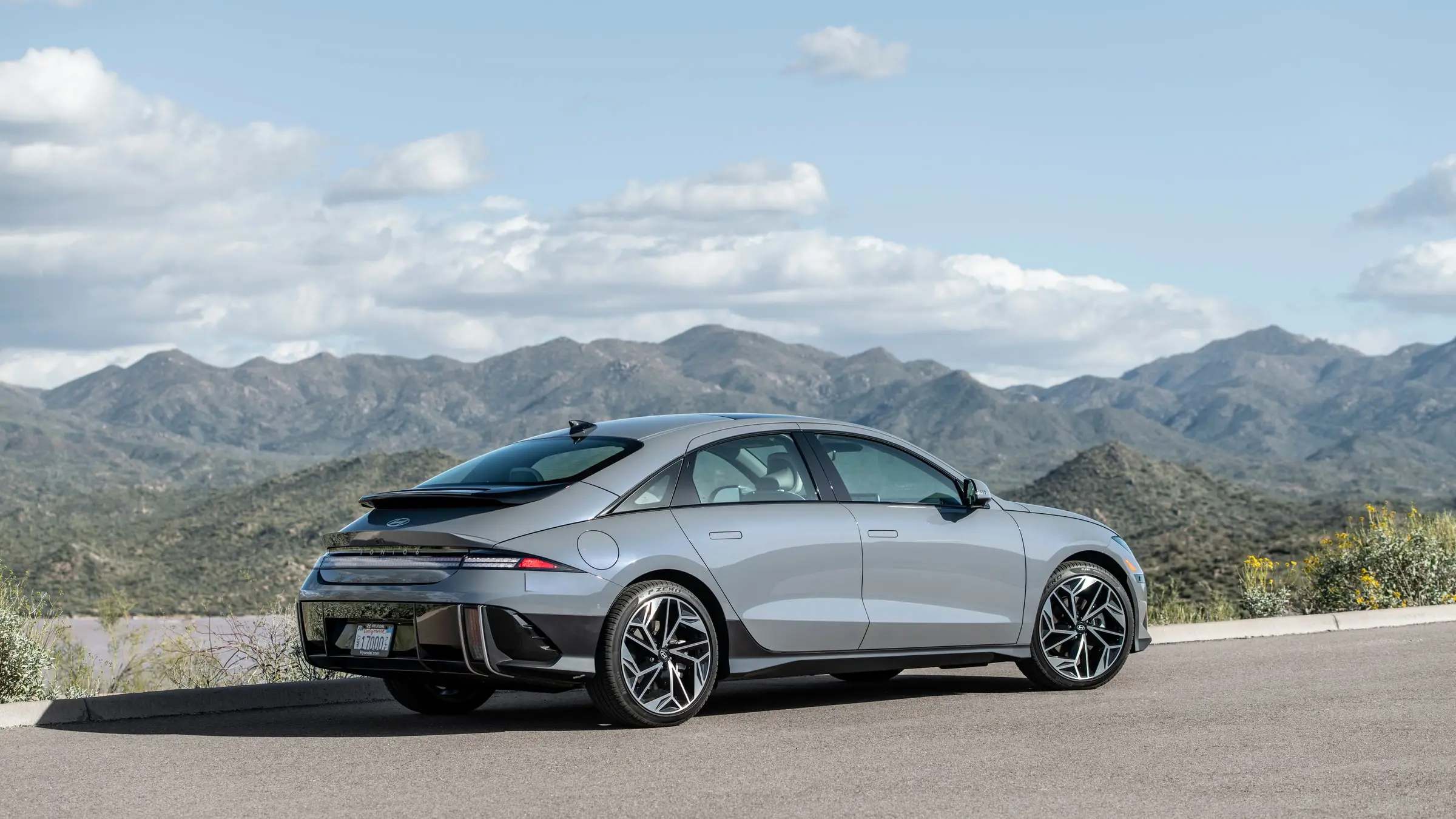 A silver-grey Hyundai Ioniq 6 Limited seen from the rear three-quarter, parked on a paved overlook with green desert mountains behind it