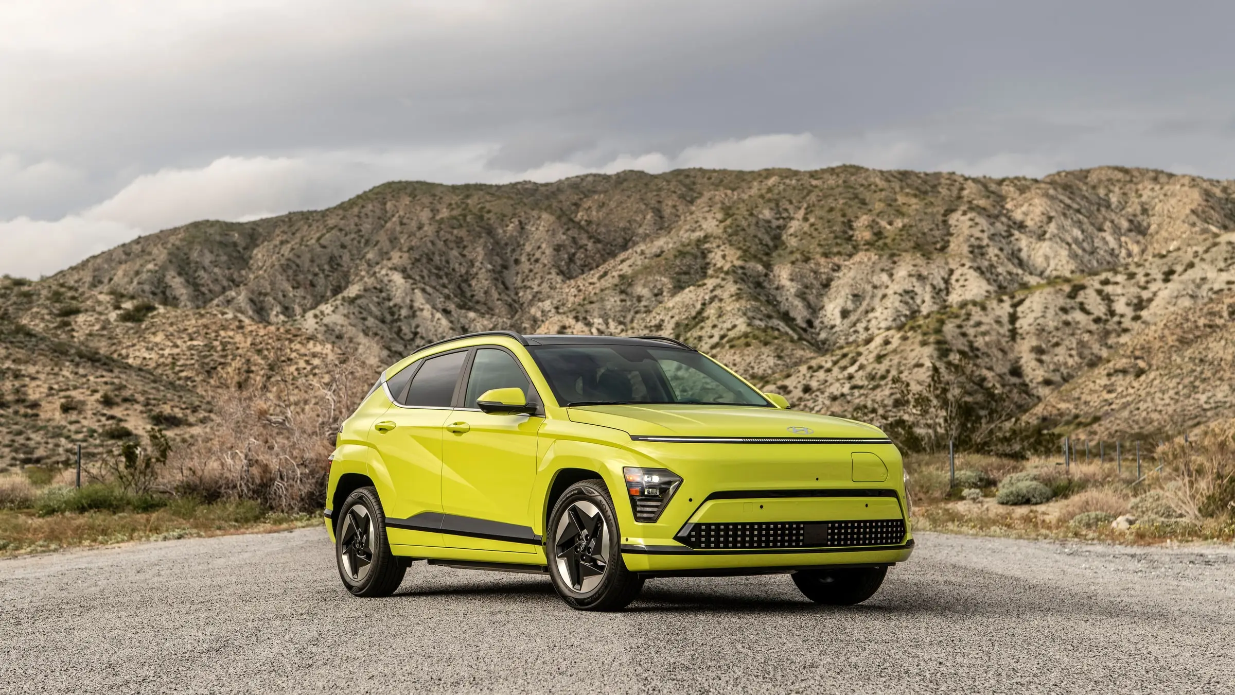 A yellow-green Hyundai Kona Electric with a black roof parked on gravel in front of dry desert mountains under a cloudy sky