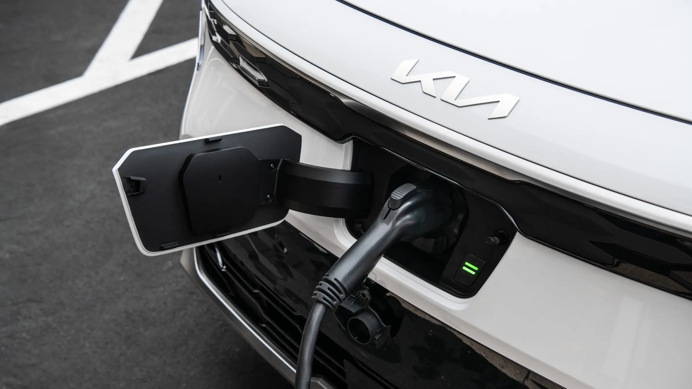A charging cable plugged into the open charge-port door in the nose of a white Kia Niro EV, below the Kia wordmark