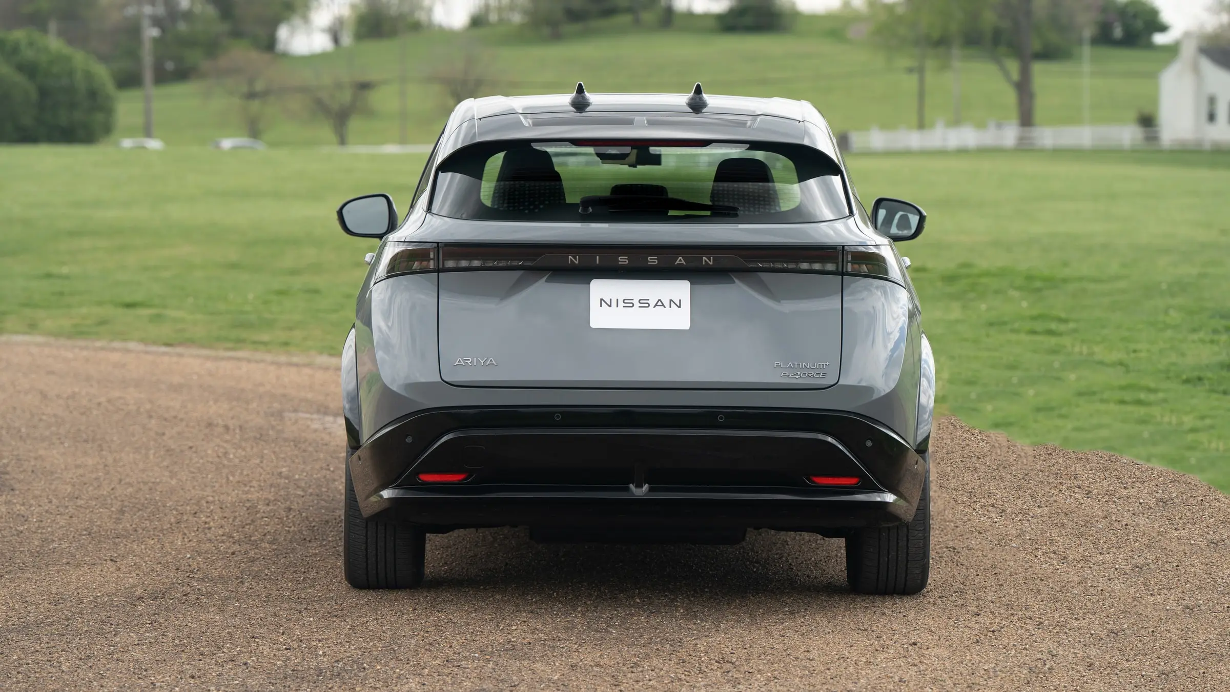 A grey Nissan Ariya Platinum+ e-4ORCE seen from directly behind, parked on a gravel drive beside a green pasture with a white fence