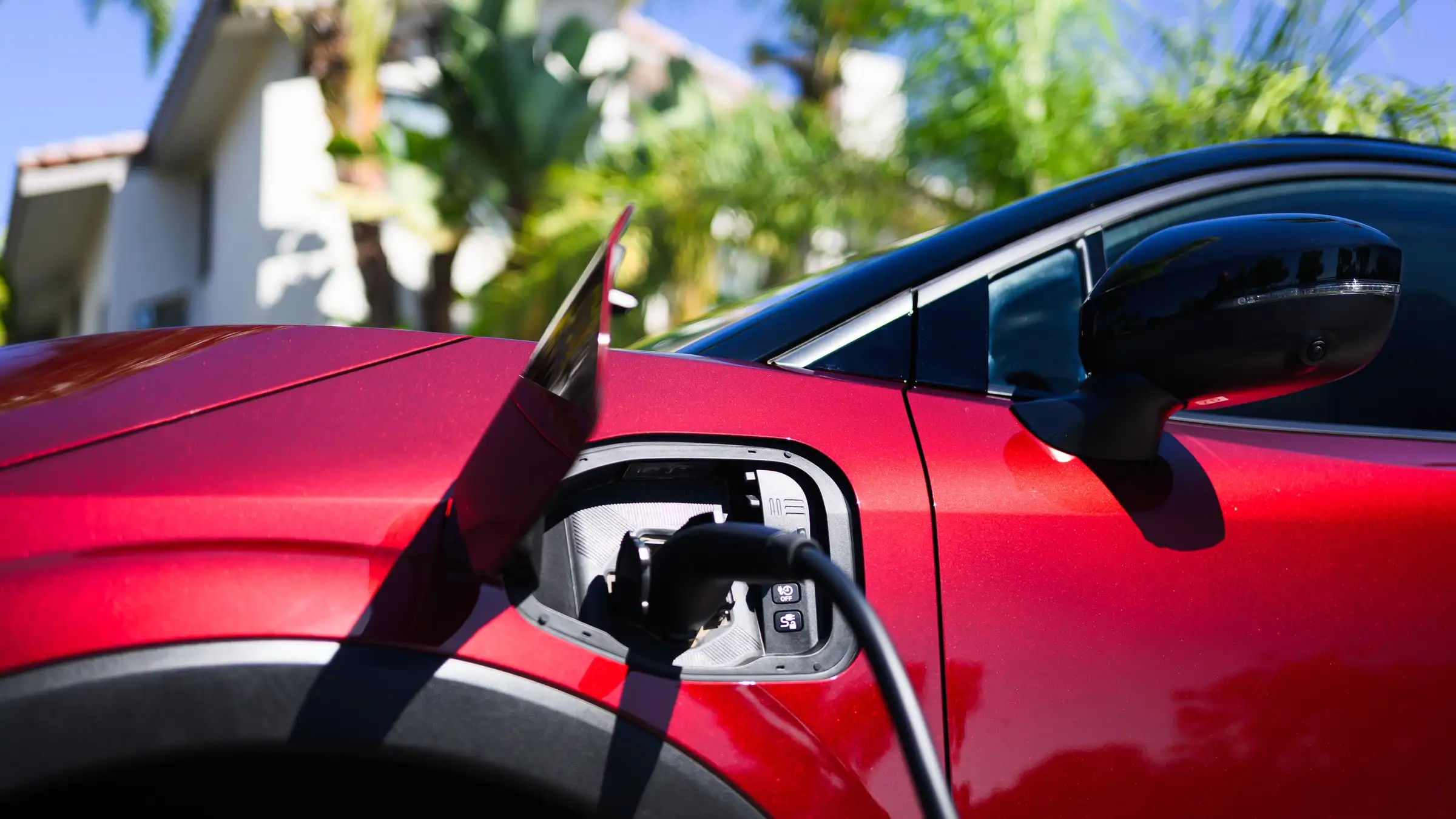 A red electric car plugged in at home, its charge door open, with a tile-roofed house and palm trees behind it