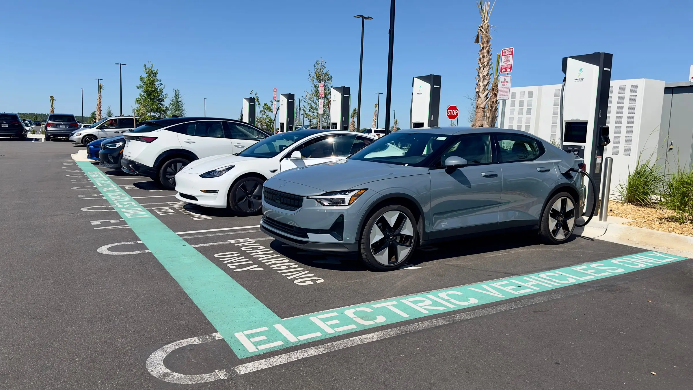 A grey Polestar 2 plugged in at an Electrify America fast charger in a Costco parking lot, beside a white Tesla Model 3 under a clear blue sky