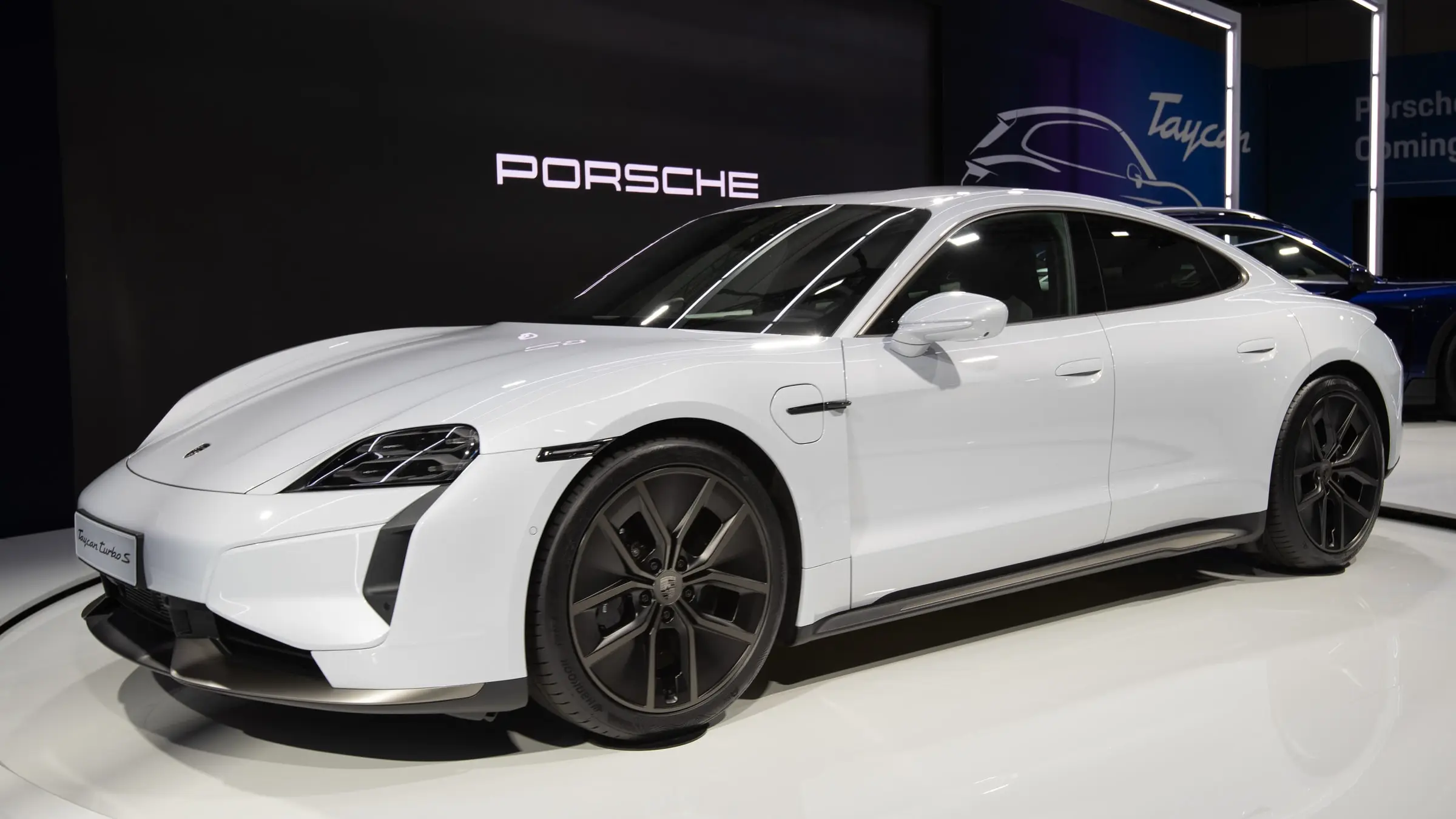 A white Porsche Taycan Turbo S on an auto show turntable in front of a Porsche and Taycan backdrop
