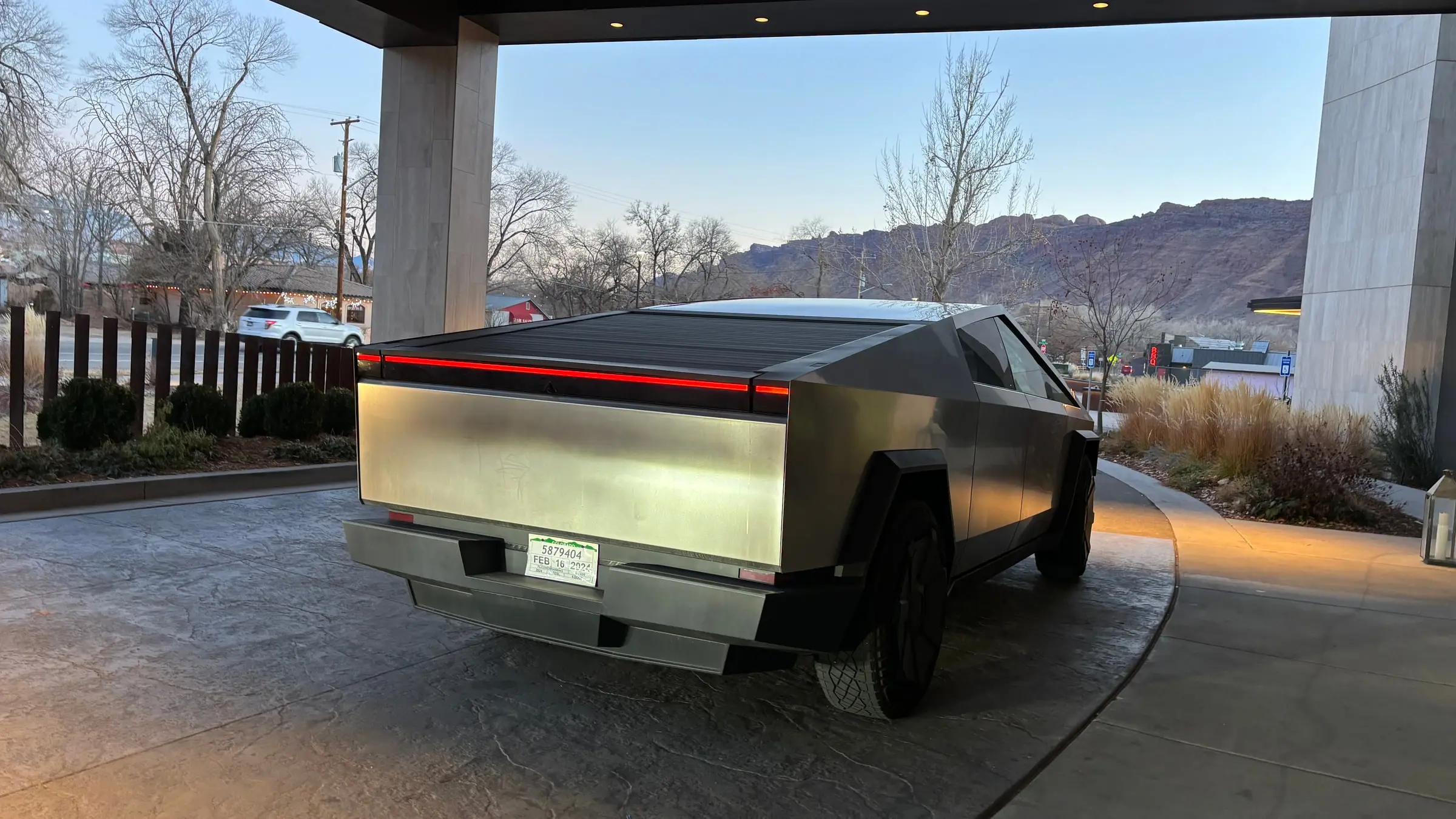 A stainless-steel Tesla Cybertruck parked under an entrance canopy at dusk, with red rock cliffs beyond