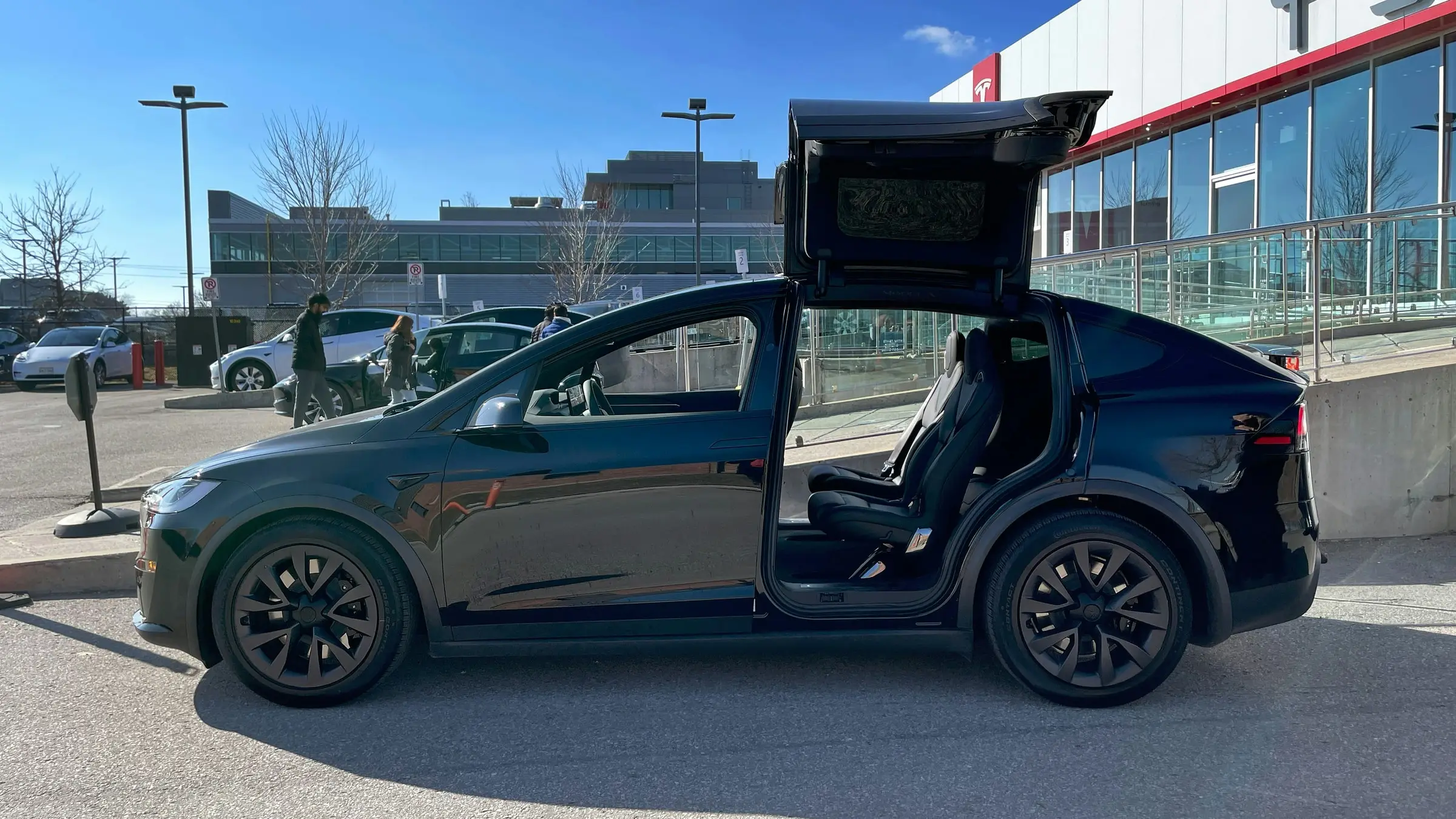 A black Tesla Model X with its rear falcon-wing door raised, parked outside a Tesla store under a clear sky