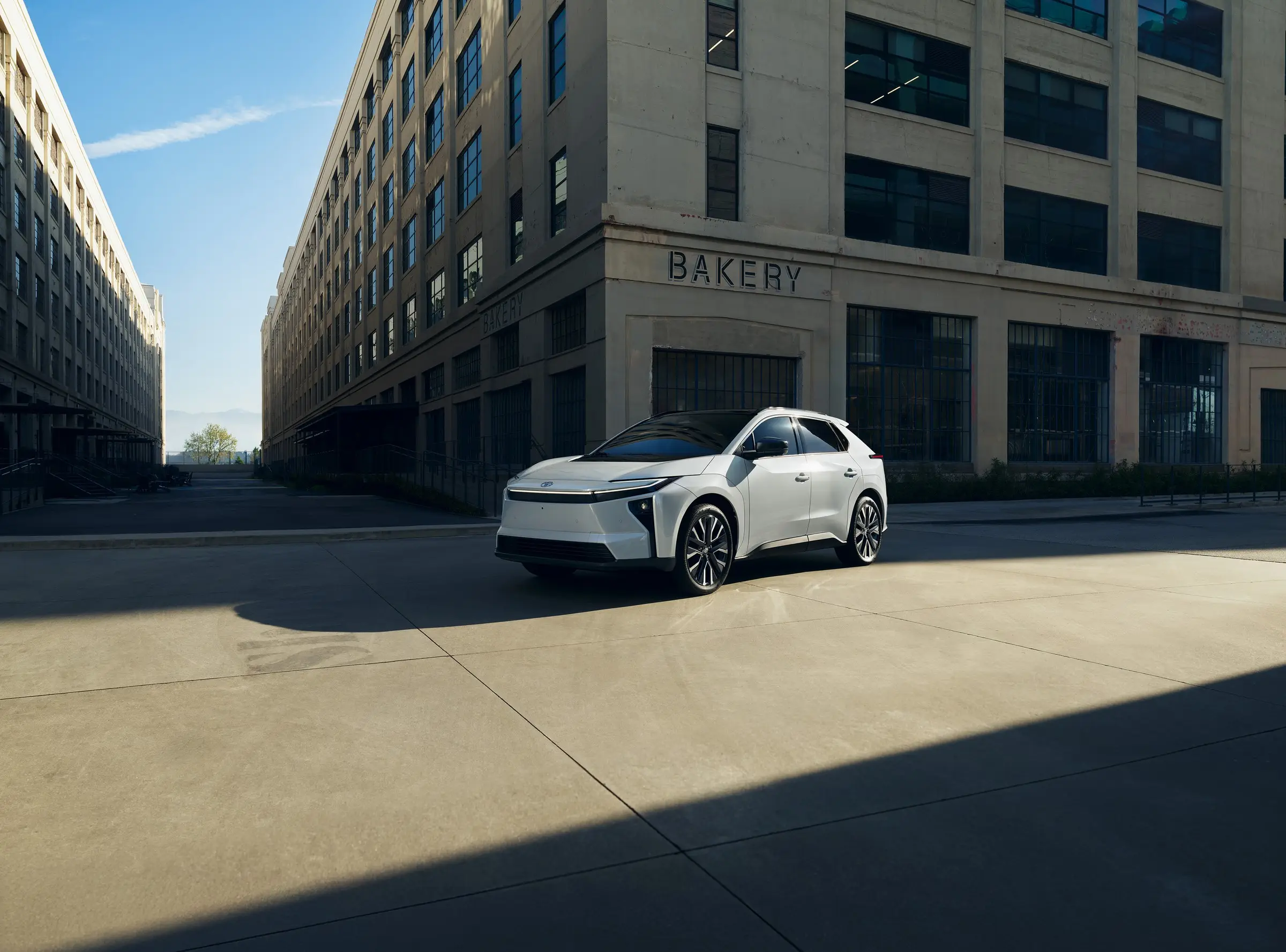 A white Toyota bZ parked on a sunlit street between industrial buildings